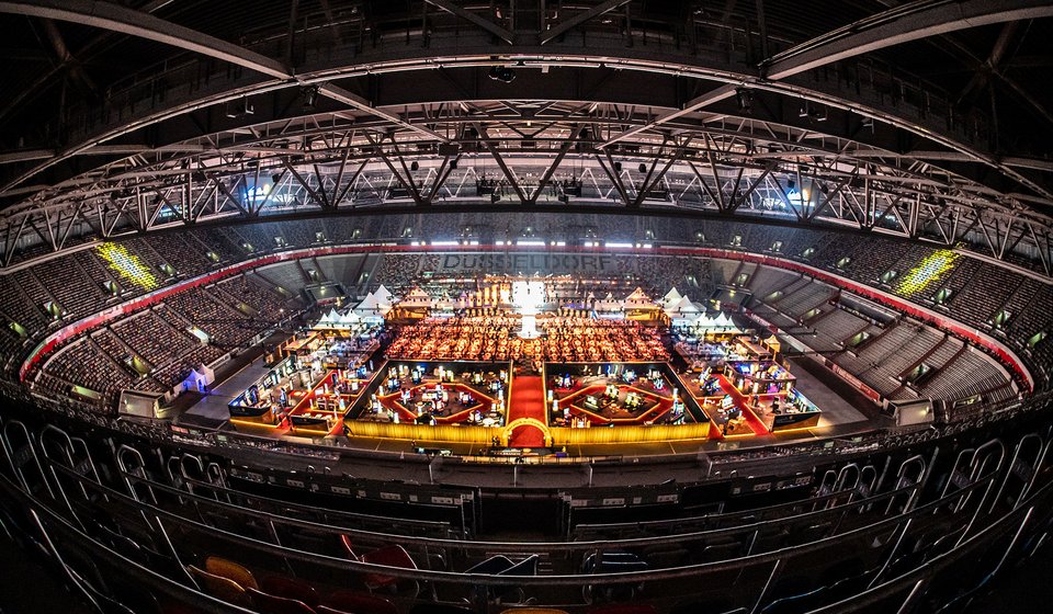 Das Foto zeigt das Innere der Merkur Spiel-Arena in Düsseldorf während eines Events. Die Arena ist weitläufig und von zahlreichen Sitzreihen umgeben, die sich bis zum Dach hinaufziehen. Das Zentrum ist in ein lebhaftes Veranstaltungsgelände umgewandelt, mit mehreren hell erleuchteten und farbig markierten Ständen, die wie eine Messe oder ein Markt aussehen. Die strukturelle Architektur des Stadions, mit freiliegenden Trägern und einem komplexen Dachwerk, ist deutlich sichtbar. Das Licht in der Halle ist gedämpft, was den Blick auf die lebendige Aktivität unten auf dem Boden lenkt, während die Sitzplätze größtenteils leer zu sein scheinen. Über dem Eventbereich sind die Worte "DÜSSELDORF" in großen Buchstaben erkennbar, die die Lokalität hervorheben. Das Foto fängt die Größe und die Atmosphäre der multifunktionalen Veranstaltungsstätte ein.
