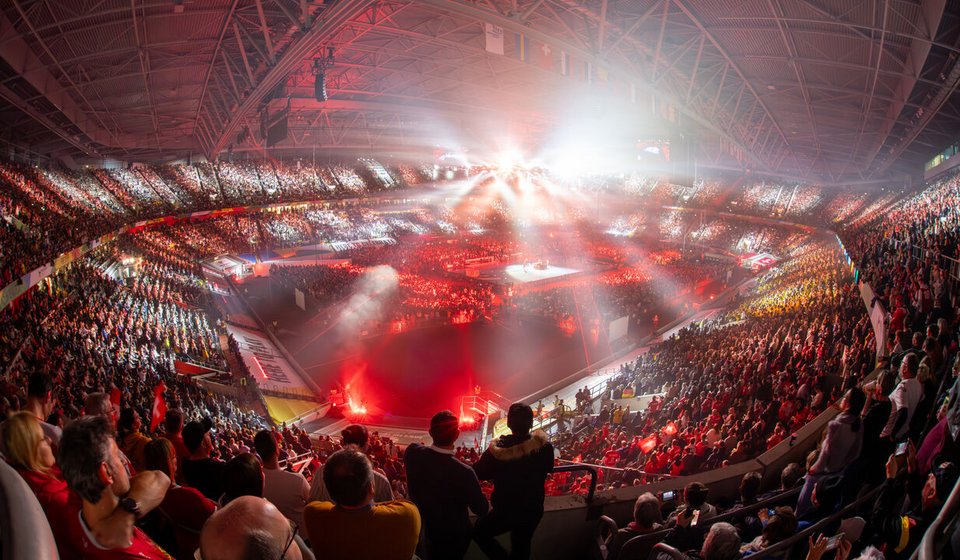 Das Bild zeigt eine aufregende Innenansicht der Merkur Spiel-Arena während der Eröffnungsfeier der EHF EURO 2024. Die Arena ist mit einer jubelnden Menge gefüllt, deren Energie durch die rot und warmen Lichter verstärkt wird, die über das gesamte Stadion verteilt sind. Im Zentrum befindet das beleuchtete Handballspielfeld, das den Blickfang der Zuschauer bildet. Die Dachkonstruktion und die Beleuchtung der Arena erzeugen eine dramatische und festliche Atmosphäre, die perfekt zu dem sportlichen Großereignis passt.