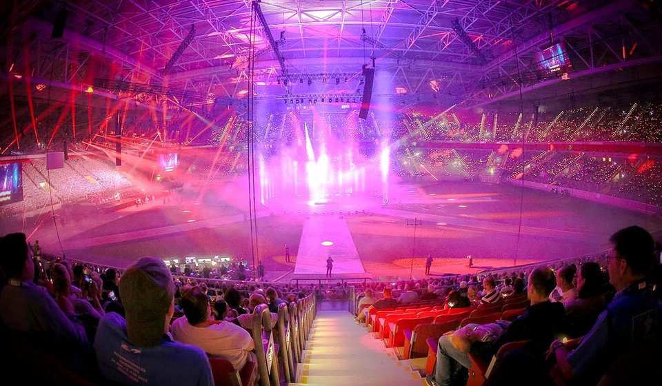 Das Bild zeigt eine beeindruckende Innenaufnahme der Merkur Spiel-Arena während eines Pop-Oratoriums. Das Stadion ist erfüllt von einem weichen, rosafarbenen Schein, der durch die Bühnenlichter erzeugt wird und sich im gesamten Raum ausbreitet. Die Zuschauer sitzen in leuchtend roten Sitzen und blicken auf die Bühne, wo die Performance stattfindet. Die Bühne selbst ist ein Zentrum intensiver Lichtstrahlen, die nach oben in die Stadionstruktur und in die Menge richten. Überall im Raum funkeln Lichtpunkte, was darauf hindeutet, dass die Zuschauer Lichtquellen oder Handys halten, um den Moment festzuhalten. Die Perspektive des Fotos suggeriert, dass der Betrachter selbst Teil des Publikums ist, und fängt die Größe des Ereignisses und die Faszination der anwesenden Zuschauer ein.