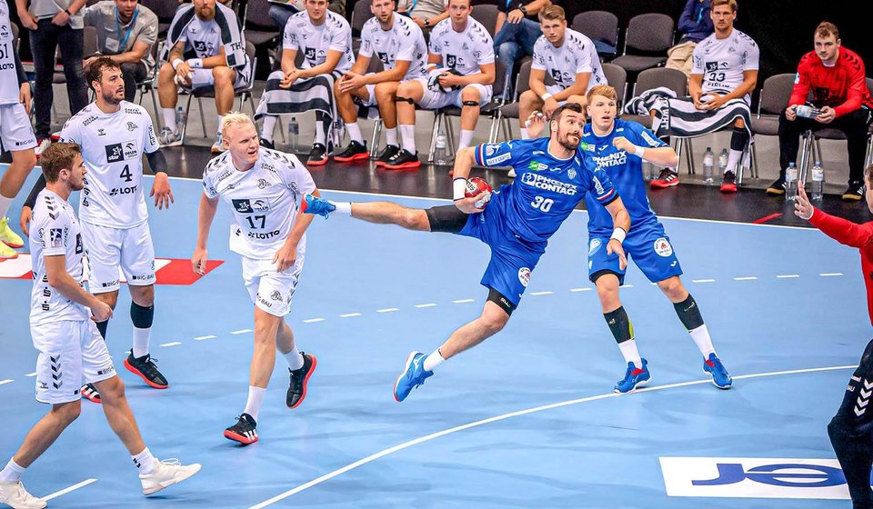 Handballer setzt zum Wurf auf das Tor an. Um ihn herum sind sowohl Spieler aus seiner Mannschaft, als auch Spieler aus der gegnerischen Mannschaft. 