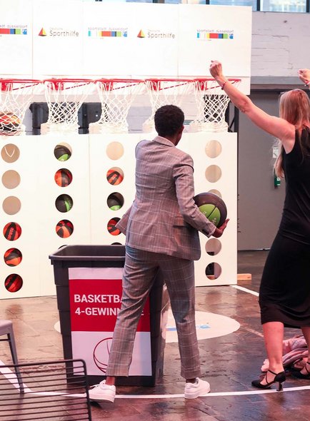 Vier Personen stehen vor einer Basketball-4-Gewinnt Wand. Eine Frau im langen Kleid jubelt, während sich ein Mann im Anzug für den Wurf bereitmacht. 